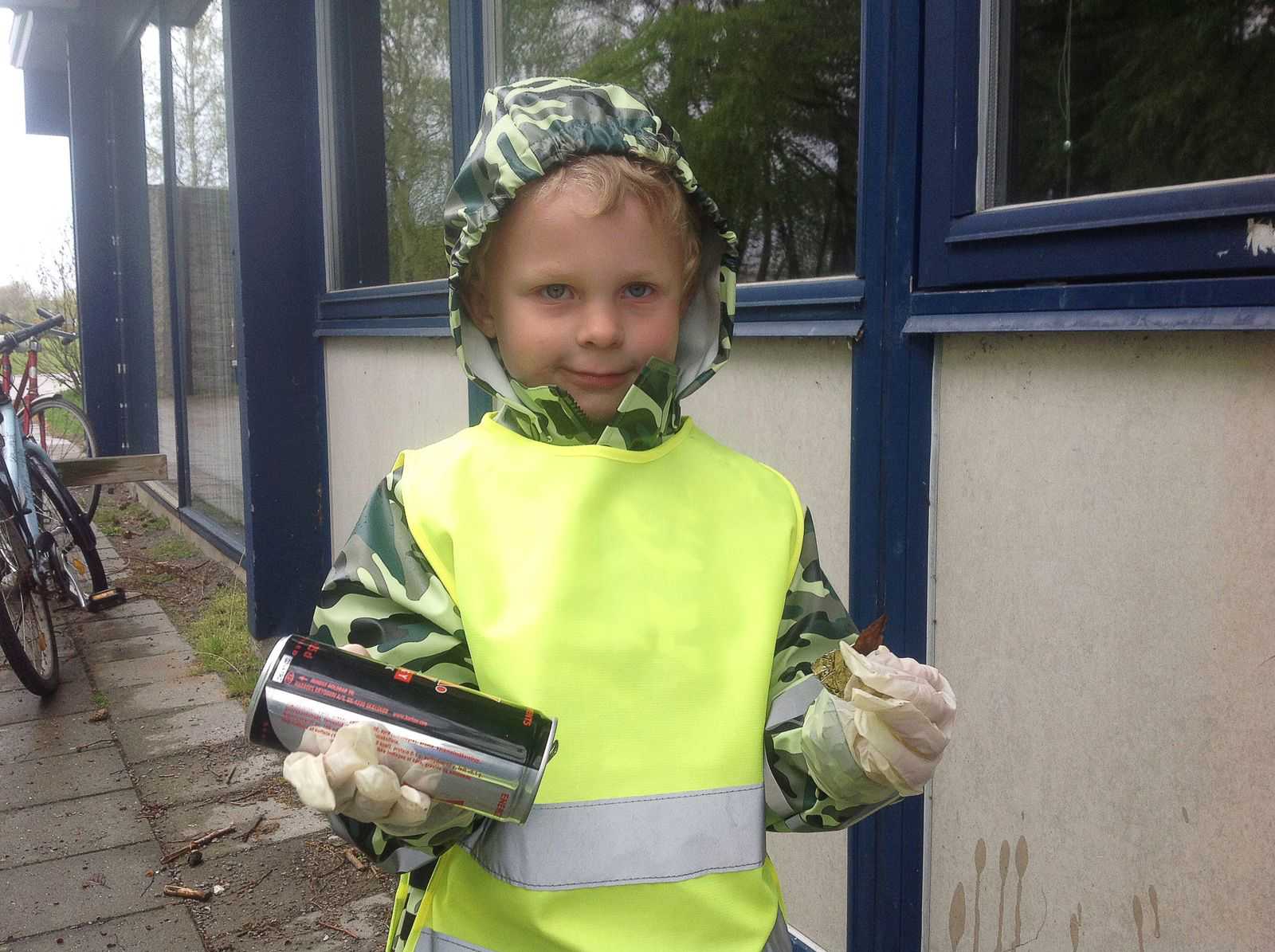 Lille dreng iført camouflagefarvet regntøj, gul vest og gummihandsker holder en dåse i den ene hånd og et stykke affald i den anden