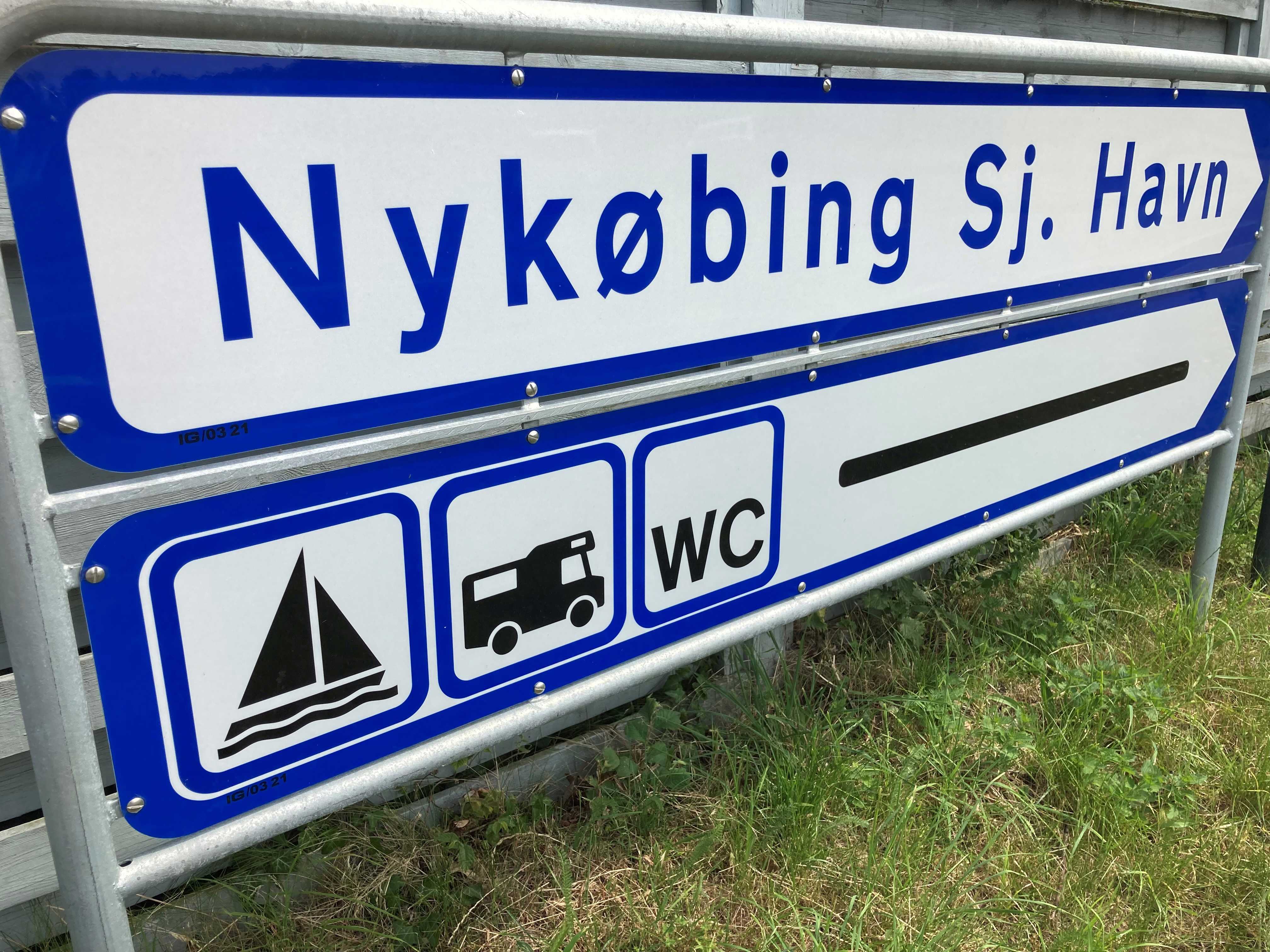 Blåt/hvidt vejskilt med teksten "Nykøbing Sjælland Havn" og ikoner for båd, autocamper og WC