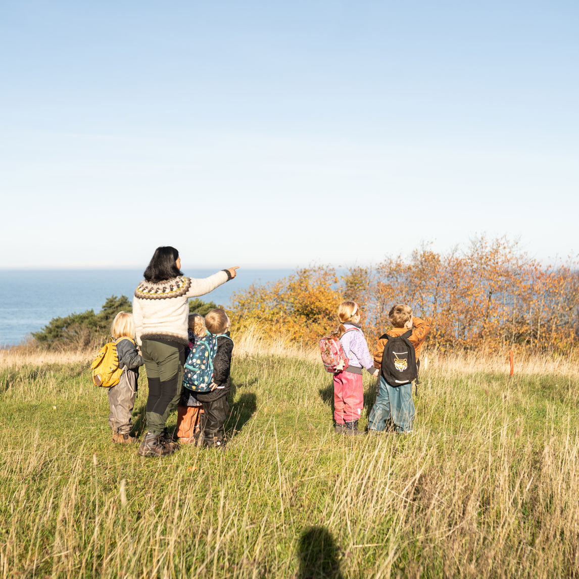 En voksen og flere børnehavebørn der står og ser ud over havet