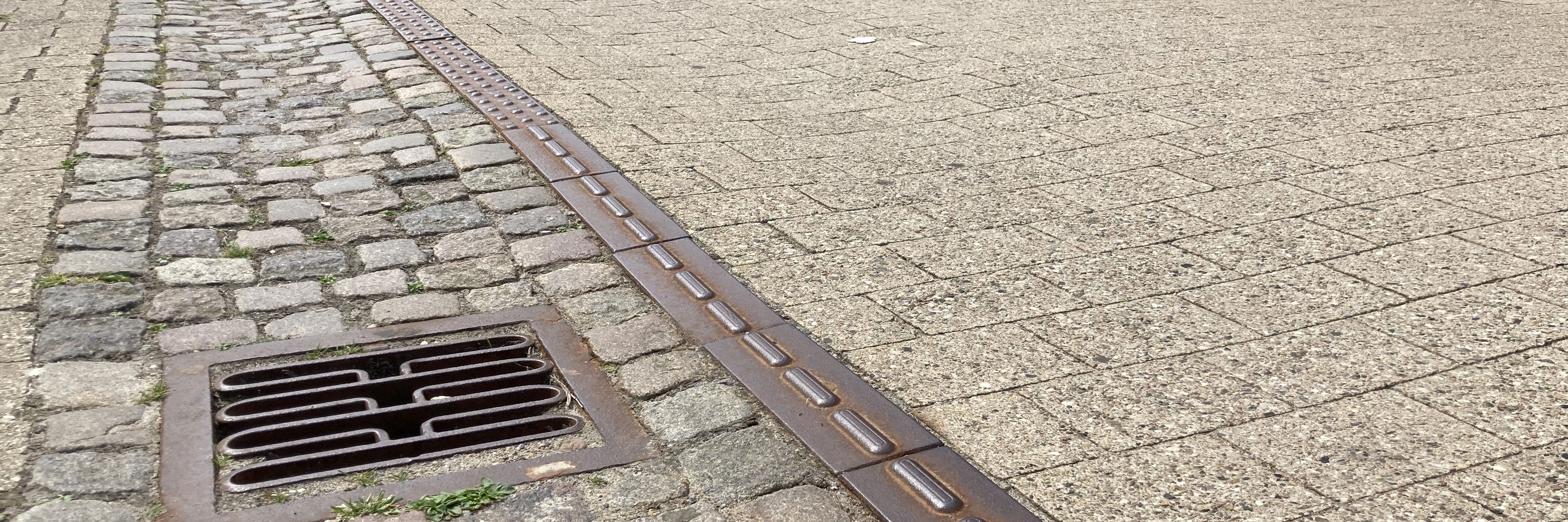 Closeup af brostensbelægningen i Algade, hvor gågaden er i fokus og fremstår nyfejet. I baggrunden fornemmer man handelslivet og butikkerne