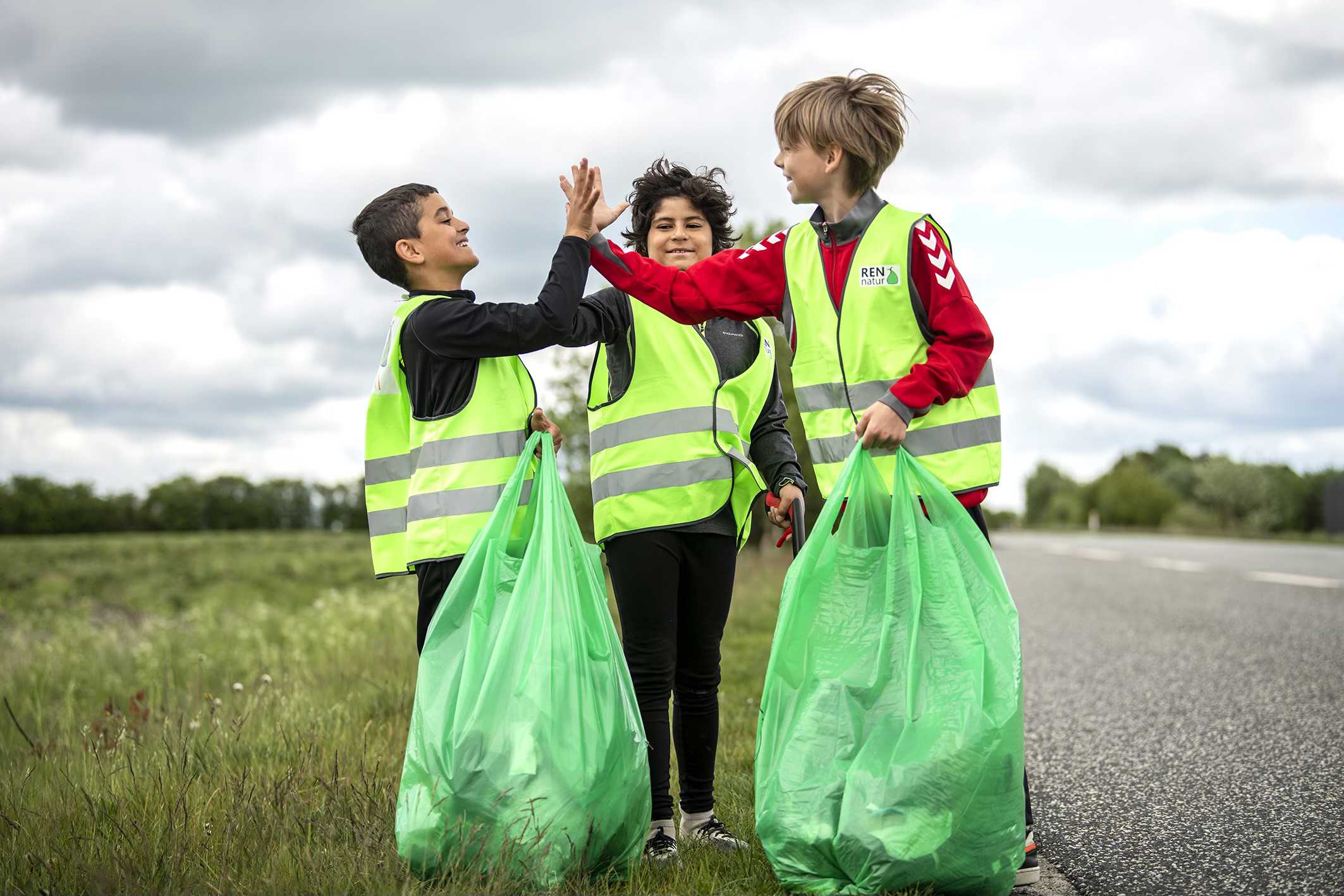 3 drenge iført gule veste og grønne affaldssække smiler og giver high five i en grøn græsrabat