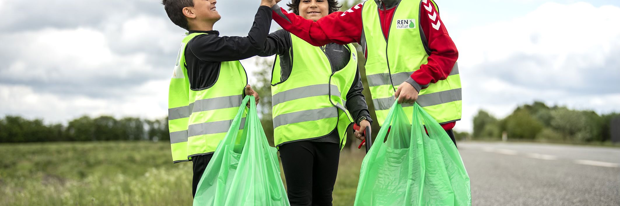 3 drenge iført gule veste og grønne affaldssække smiler og giver high five i en grøn græsrabat