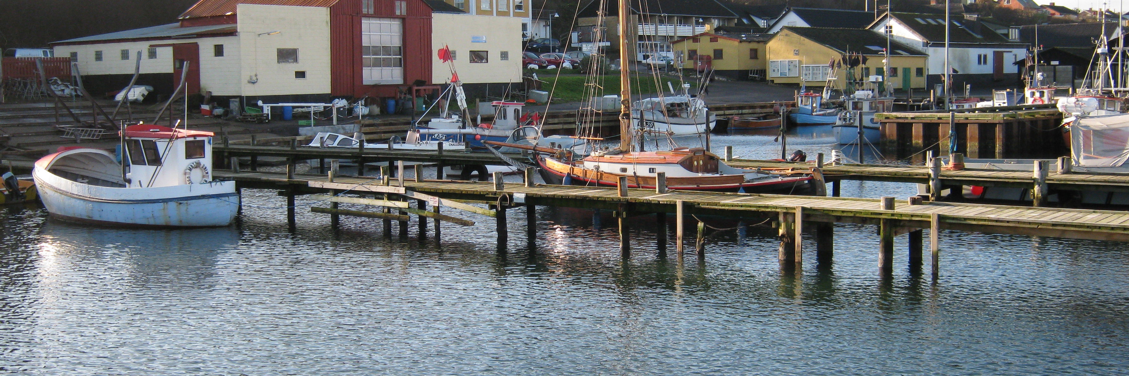 Kig fra vandet mod havnefronten. I forgrunden moler og både. I baggrunden huse på havnen.