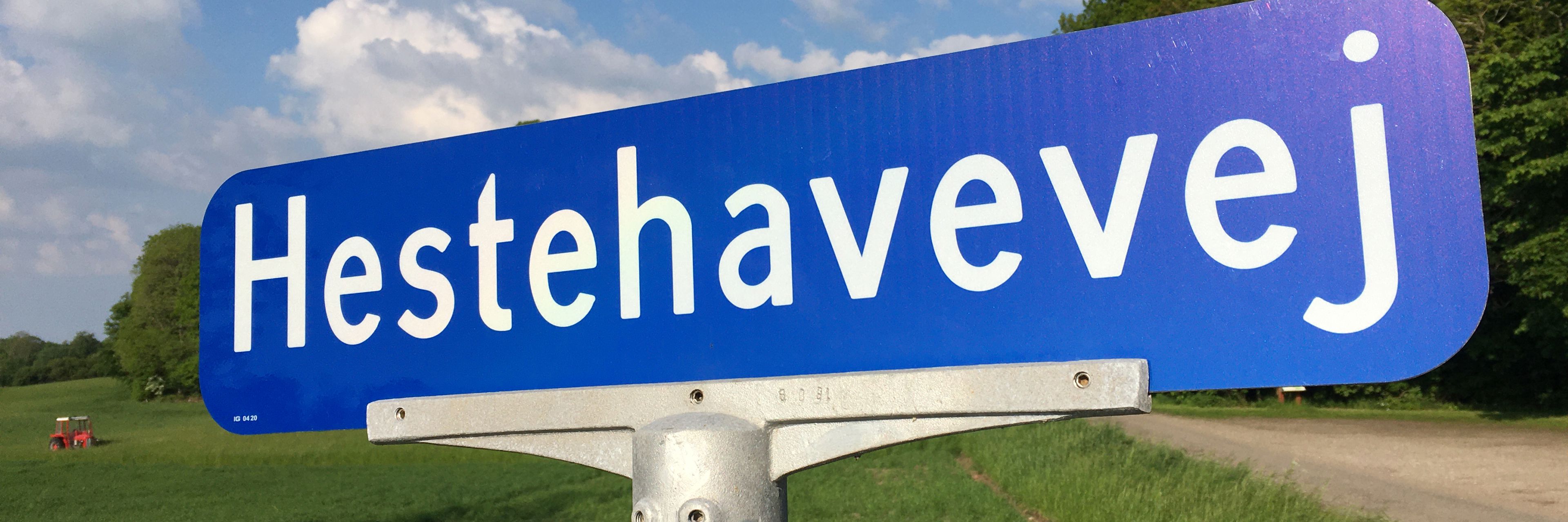 Blåt vejskilt med vejnavnet "Hestehavevej" - i baggrunden ses en mark med en lille rød traktor